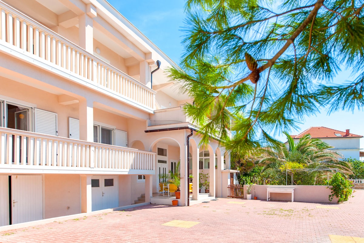 The exterior of the apartment showcases a spacious patio area paved with tiles and bordered by low shrubs. A two-story building is visible, featuring balconies with railings and large windows. Surrounding greenery adds a natural element to the inviting entrance.