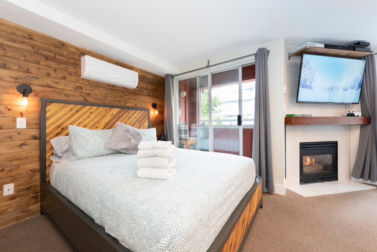 A cozy bedroom features a spacious bed with a textured duvet, glowing lights on either side, and a gas fireplace. Large windows allow natural light and provide access to the balcony, while a wall-mounted TV adds modern convenience.