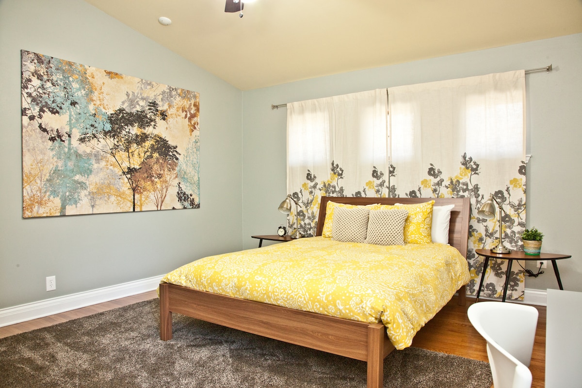 A bright bedroom features a spacious bed dressed in a yellow floral duvet. Two bedside tables hold lamps on either side. A large artwork of trees adorns the wall above the bed, complemented by soft, sheer curtains filtering natural light.