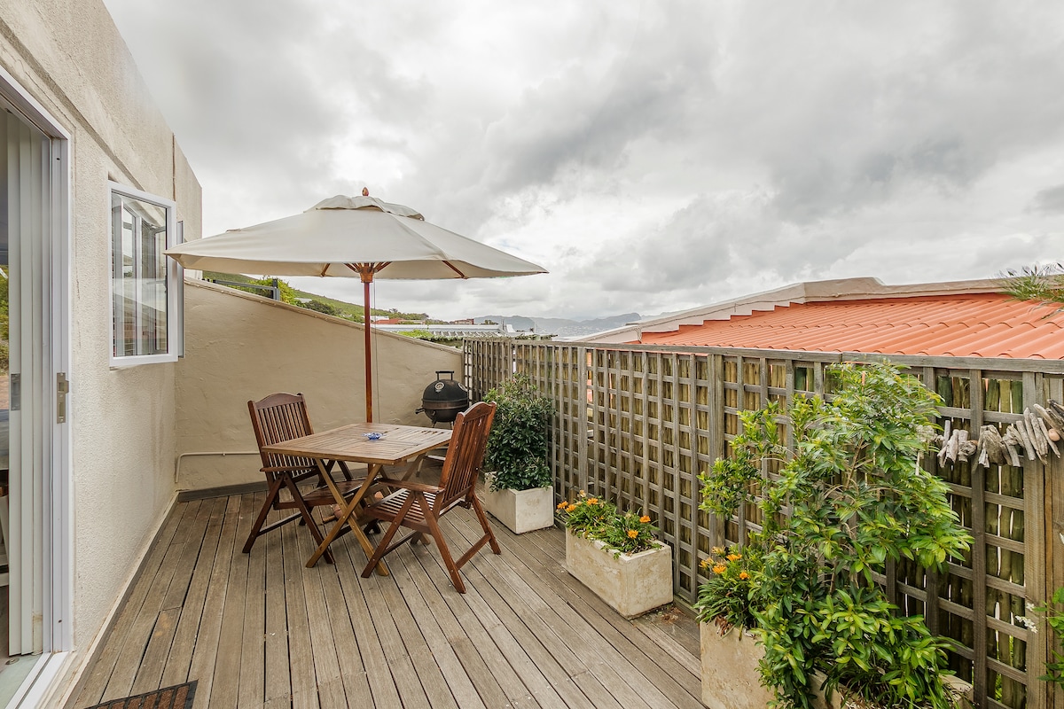 A wooden deck area is presented, featuring a round umbrella protecting outdoor seating. A table and chairs are visible, accompanied by potted plants with vibrant foliage. A barbecue grill sits nearby, and cloudy skies provide a soft, diffused light over the scene.