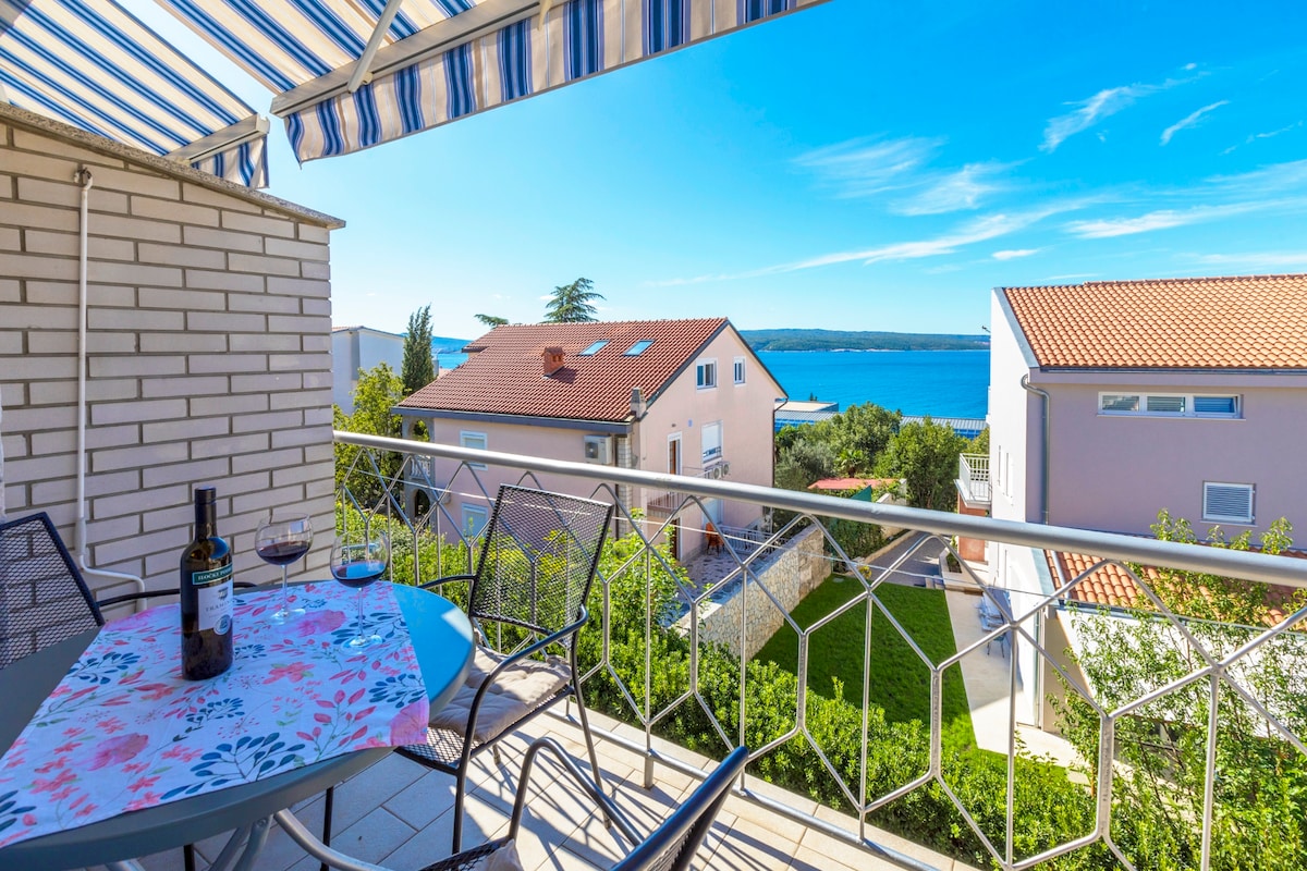A private balcony is visible, featuring a small table set for two with a floral tablecloth and wine glasses. A railing offers a clear view of the sea and neighboring properties, complemented by bright blue sky and scattered clouds. The terrace is partly covered, providing shade.