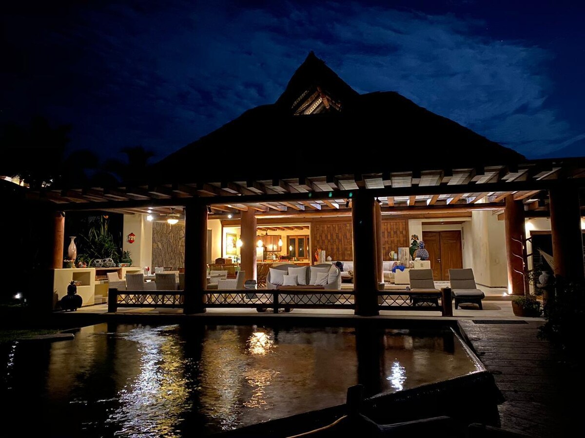A luxurious villa is illuminated under the night sky, showcasing a spacious open-air design. Large wooden columns support the thatched roof, while soft lighting highlights the inviting living area and surrounding outdoor spaces. A serene water feature reflects the tranquil ambiance.