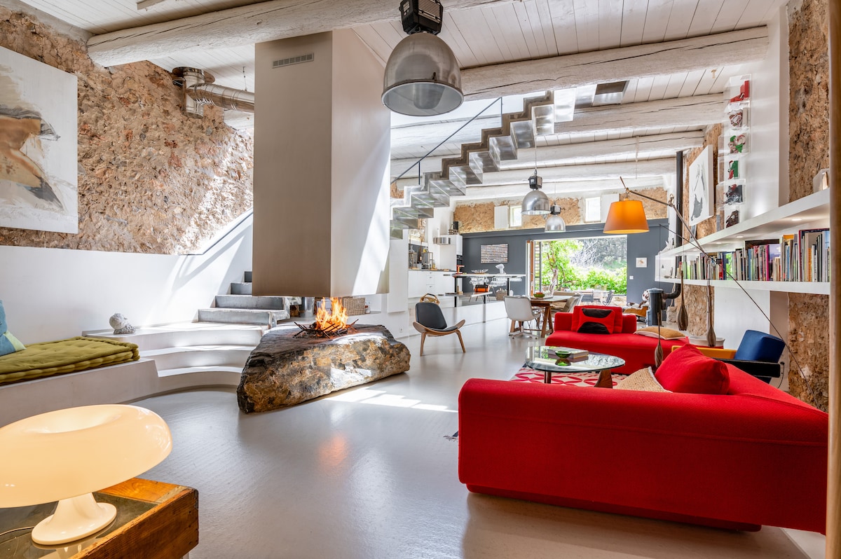 An inviting living area features a modern design with a central fireplace integrated into a natural stone formation. Bright red sofas are arranged around a low coffee table, with eclectic seating nearby. Large windows allow natural light to illuminate the space, while the unique staircase adds architectural interest.