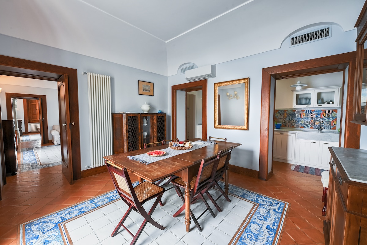 A well-lit dining area is revealed, featuring a wooden table surrounded by folding chairs. Decorative tile patterns enhance the floor, complemented by a colorful backsplash in the kitchen area. Light is provided by a ceiling fan, and a framed mirror reflects the room's warm tones.