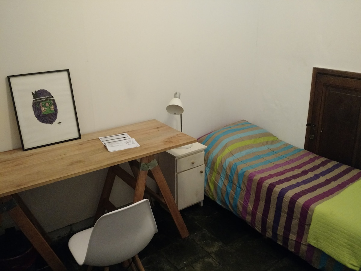 A compact room features a spacious bed draped in a striped blanket. A simple desk with a white chair is positioned beside the bed, complemented by a small lamp on the desk. A framed artwork is displayed on the wall, adding a touch of character.