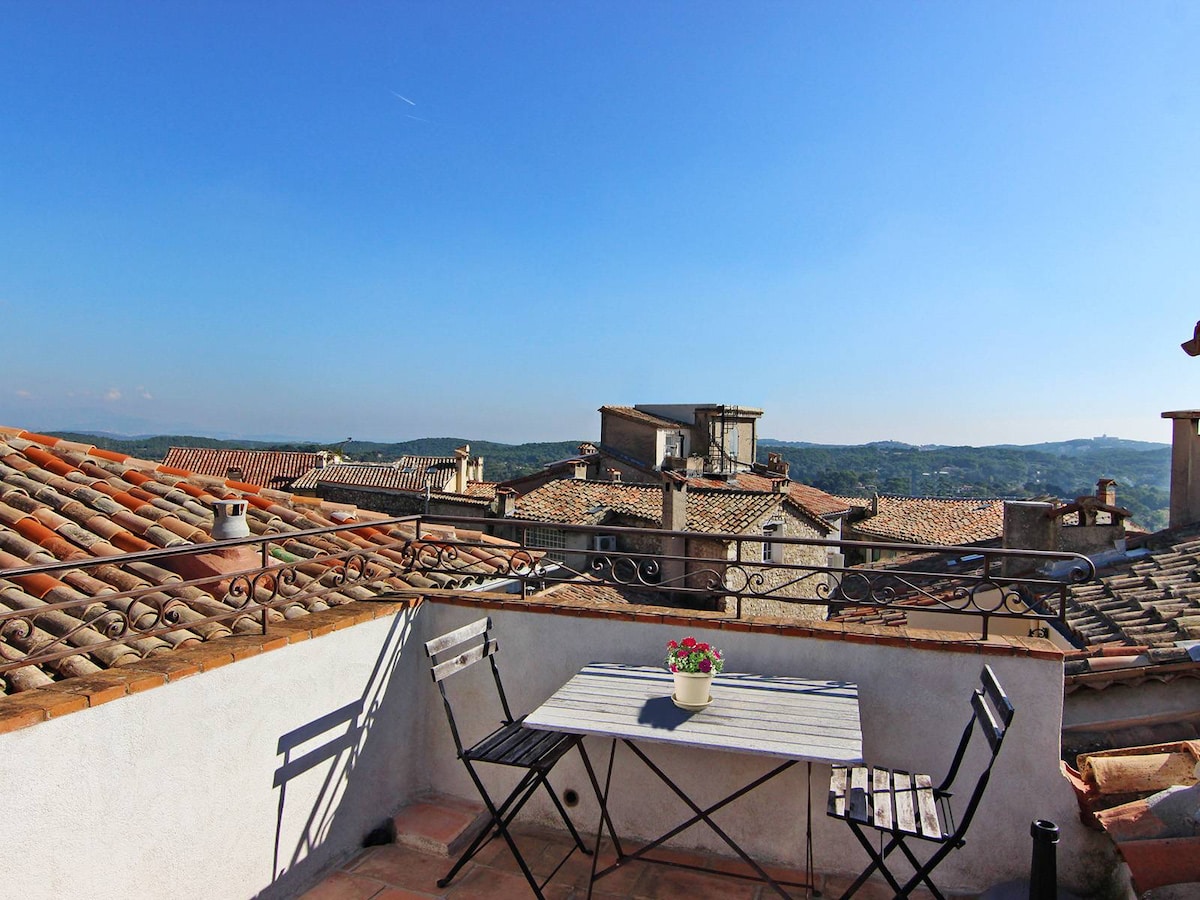 A private rooftop terrace is visible, featuring a small table and two chairs. A potted flowering plant is centered on the table. Scenic views of terracotta rooftops and rolling hills under a clear blue sky create a tranquil atmosphere.