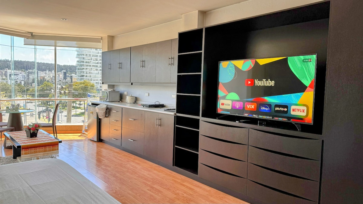A modern living space is equipped with a large Smart TV displaying various streaming options. Sleek cabinetry offers storage, and a small kitchenette is visible in the background. A window provides a view of the cityscape, filling the area with natural light.
