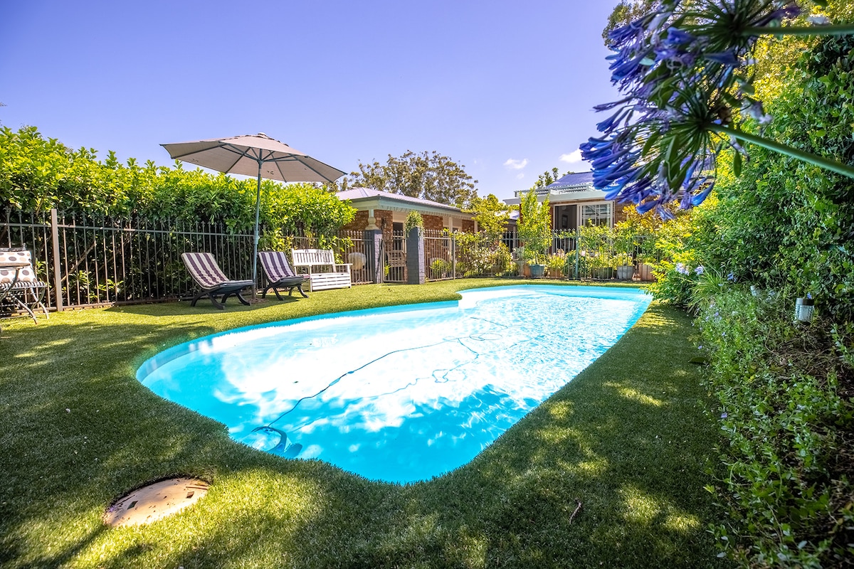 An inviting pool is surrounded by lush greenery and manicured lawns. Two lounge chairs are positioned under a large umbrella, providing shaded seating. The pool area is enclosed by a decorative fence, enhancing privacy.