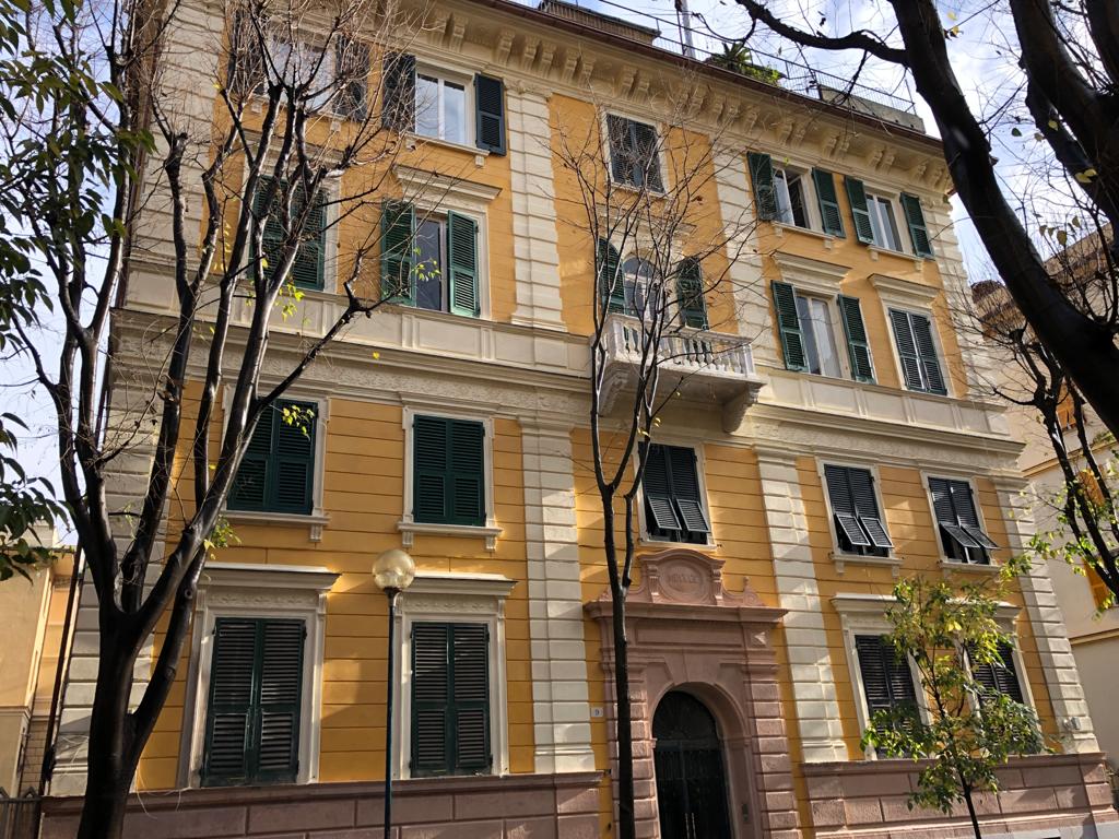 A charming building features a warm yellow facade, highlighted by white trim and green shutters. Several trees frame the exterior, enhancing the peaceful ambiance of the area. The entrance is marked by a decorative archway, inviting guests into the welcoming surroundings.