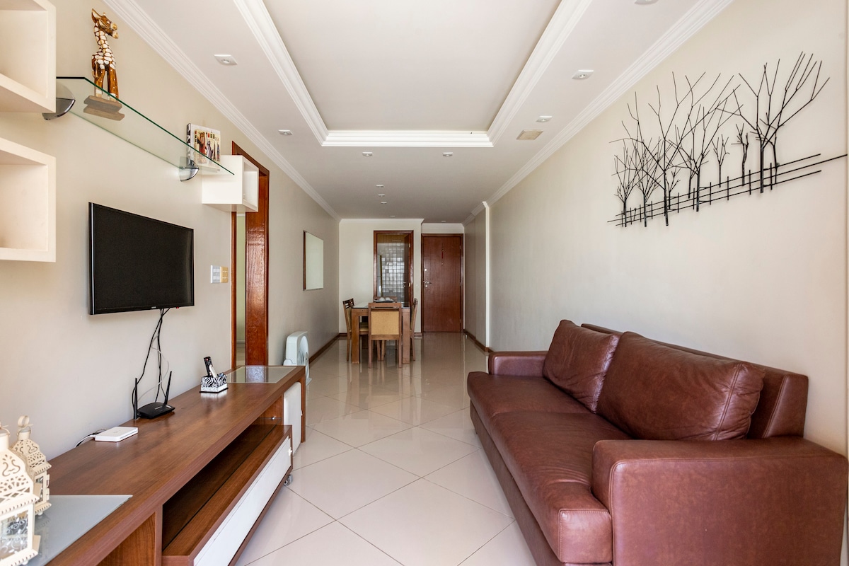 A spacious living area is observed, featuring a large brown sofa and a wall-mounted television. A wooden dining table is situated in the background, with elegant wall decor enhancing the room's aesthetic. Neutral wall colors and ample natural light create an inviting setting.