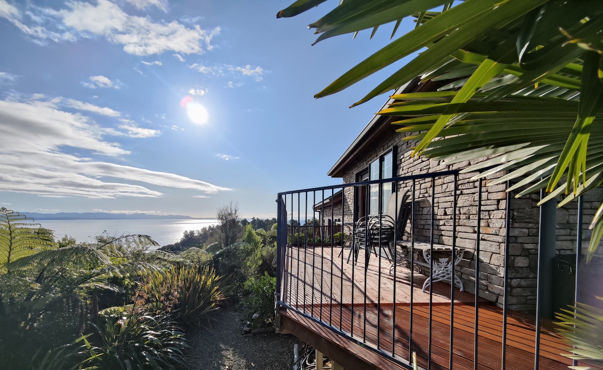 A private veranda with a wooden deck is surrounded by lush greenery and palm leaves. The scene features distant sea views under a bright sky, where sunlight reflects off the water. The exterior of the studio apartment, built with stone, complements the natural landscape.