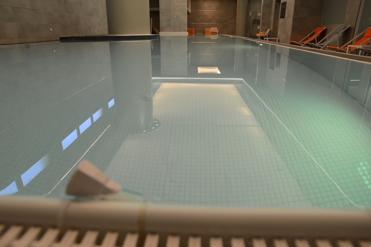 An indoor pool is lined with light blue tiles, featuring clear water that reflects soft overhead lighting. Loungers are positioned nearby, creating an inviting space for relaxation. The surrounding walls are finished in a smooth gray material, enhancing the serene atmosphere.