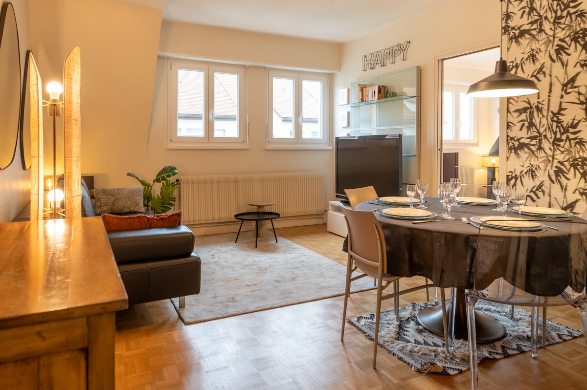 A welcoming living space features a comfortable seating area with a dark sofa and an accompanying side table. A dining table is set with glassware, surrounded by muted chairs. Natural light filters through several windows, highlighting the modern decor and wooden accents throughout.