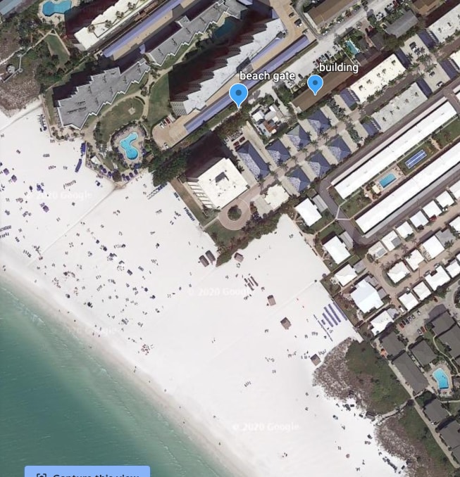 An aerial view shows a wide sandy beach bordered by the calm turquoise water. The layout includes various buildings near the beach gate, with pathways leading to the beach area. Beachgoers can be seen enjoying the shore.