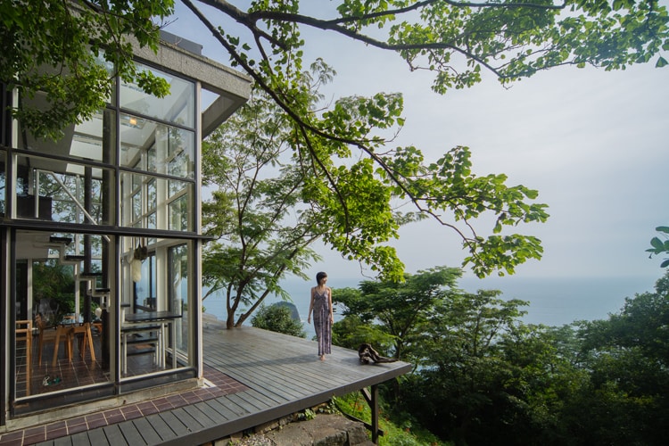 A modernist villa built with expansive glass walls is situated on a cliff surrounded by lush greenery. A large deck provides stunning ocean views. A person dressed in a patterned outfit stands on the deck, enjoying the serene atmosphere of the natural setting.