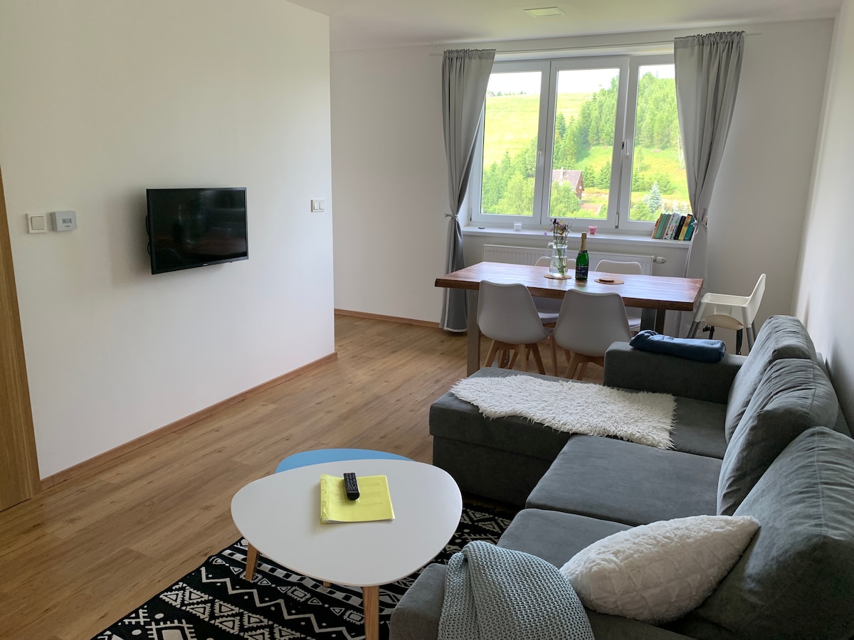 A bright living area features a gray sectional sofa and a modern coffee table adorned with a notepad. A dining table, set for four and positioned near a large window, offers views of the surrounding greenery. A wall-mounted television provides entertainment options.