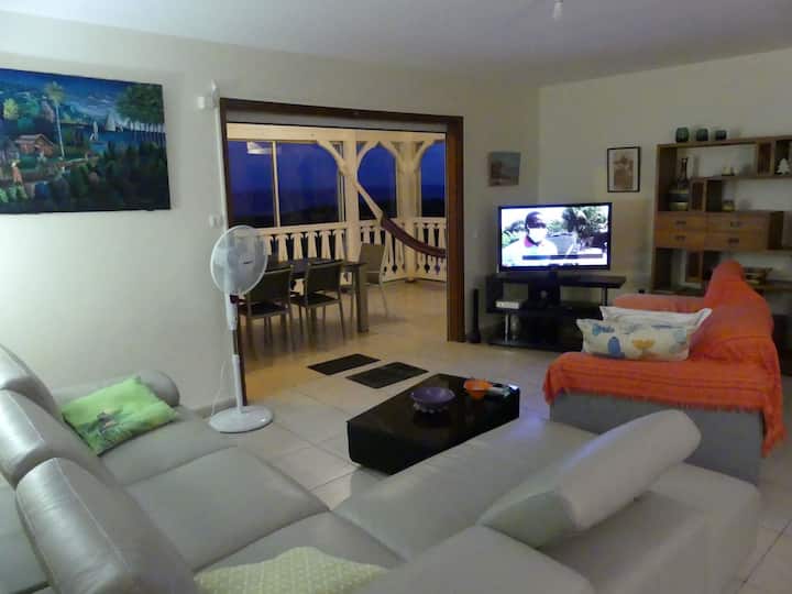 ensemble salon véranda éclairé de la villa Alizés. Au fond on aperçoit la véranda qui fait office de salle à manger. La cuisine est à droite et on traverse le salon pour servir la table.