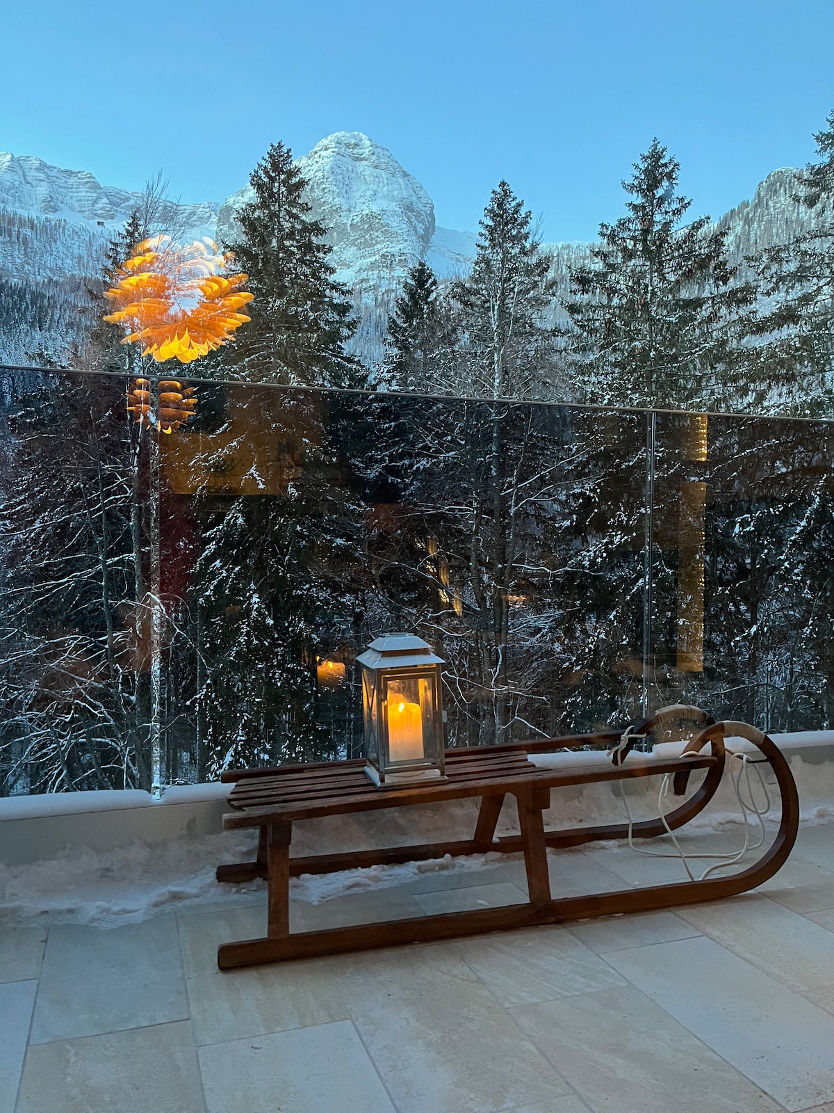 A wooden sled is positioned on a spacious balcony, accompanied by a lantern holding a candle. The backdrop features a landscape of snow-covered trees and mountains, with a warm glow from a light fixture reflecting off the glass railing.