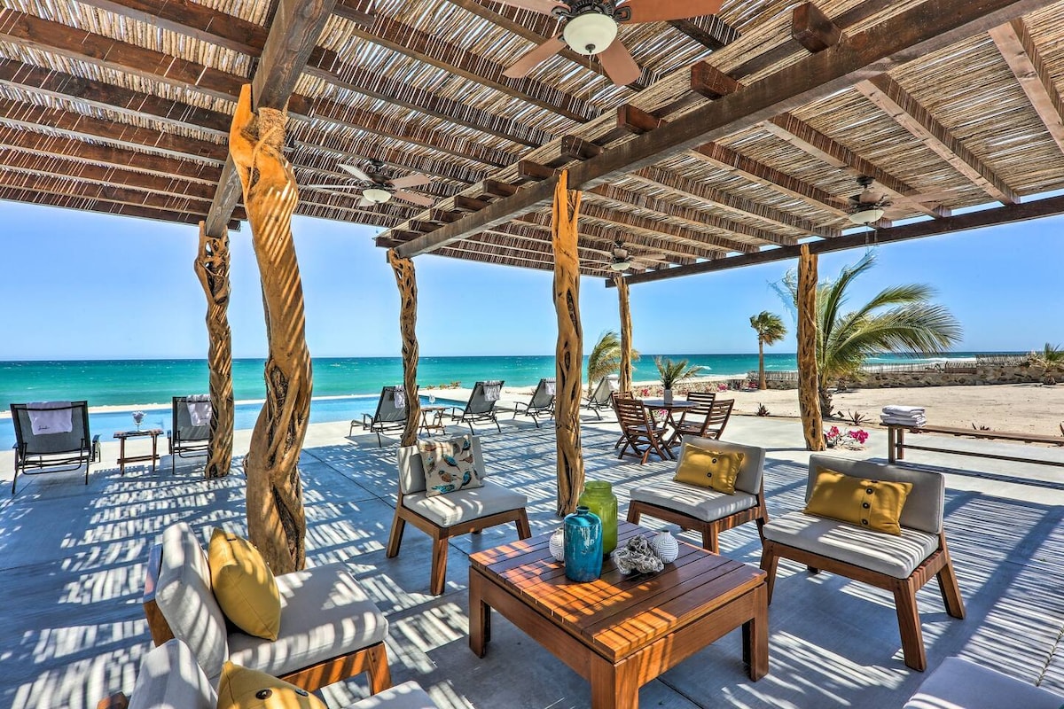 An outdoor terrace features natural wooden beams and columns, providing shade and a relaxed seating area. Comfortable sofas with cushions are arranged around a wooden coffee table, overlooking the sea. Sun loungers are positioned on the nearby patio, with palm trees framing the picturesque view.