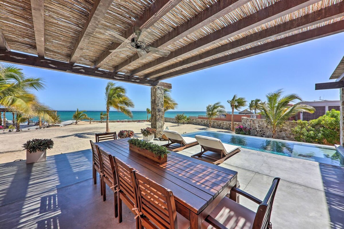 An outdoor dining area is showcased under a wooden pergola, featuring a large table surrounded by wooden chairs. In the background, an infinity pool overlooks a sandy beach and the calm sea, framed by swaying palm trees.
