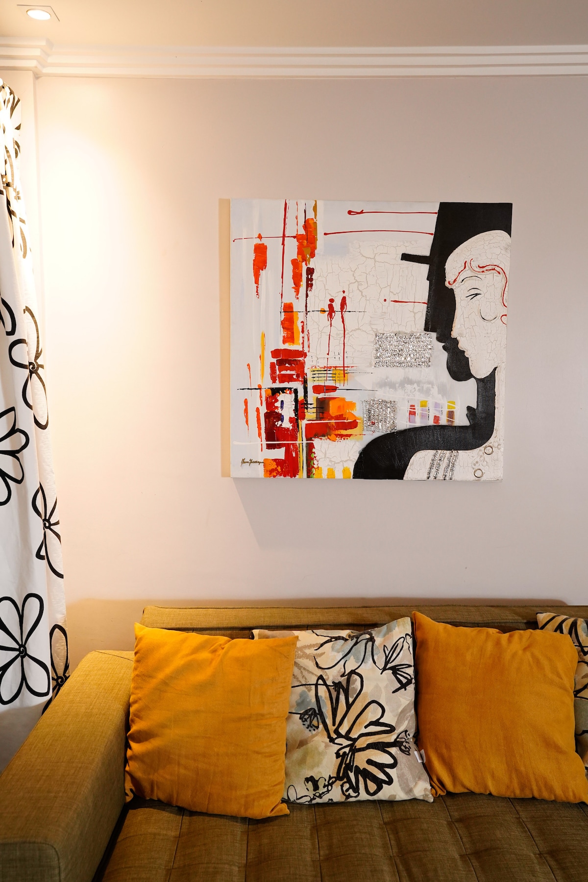 A modern art piece featuring abstract figures is displayed on a light-colored wall. Below it, a comfortable sofa adorned with decorative pillows in shades of yellow and floral patterns creates an inviting seating area.
