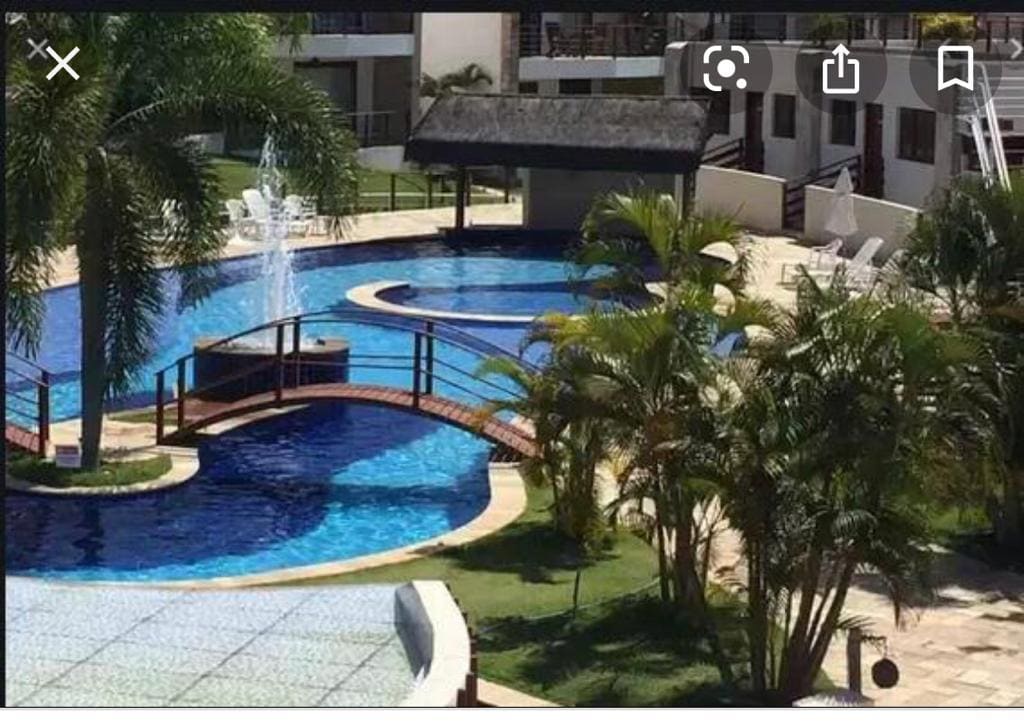 A resort-style pool area is surrounded by lush greenery and palm trees. Multiple sparkling blue pools are interconnected by wooden bridges, with a fountain adding a refreshing touch. Lounge chairs are arranged nearby, promoting a relaxed outdoor atmosphere.