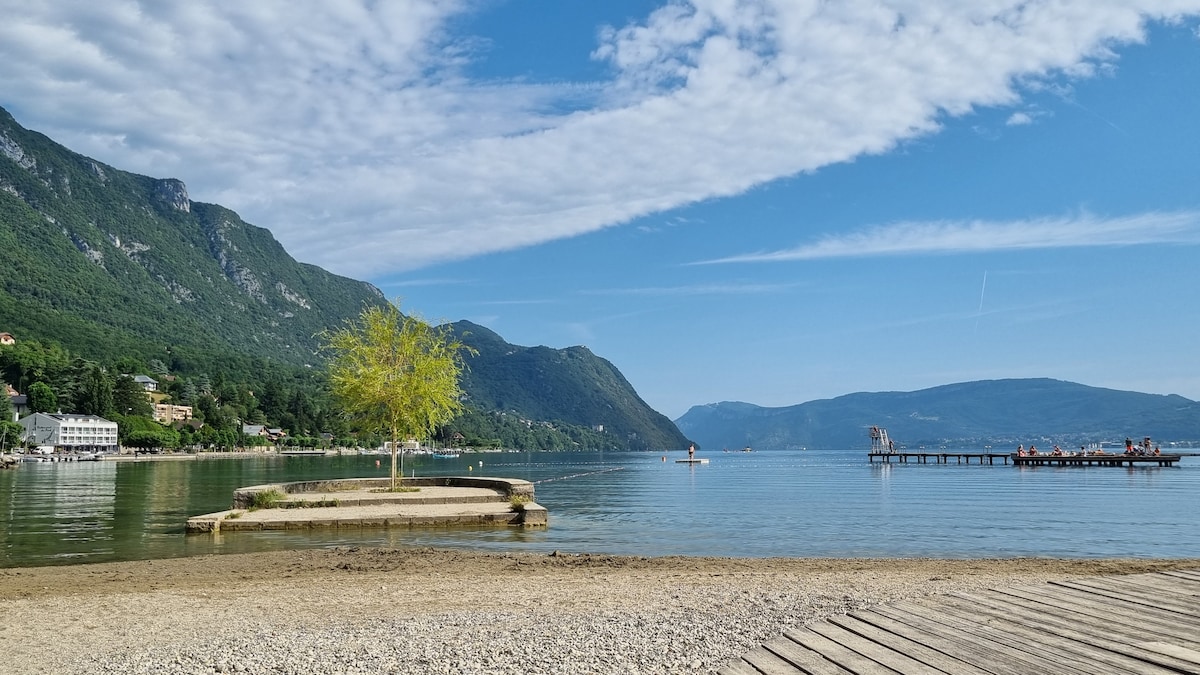 The image captures a serene lakeside view with gentle ripples on the water's surface. Mountains rise in the background, while a small island with a solitary tree is visible. A wooden pier extends into the lake, with a few boats in the distance.