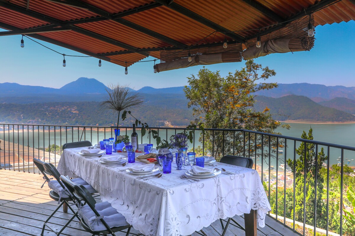 An outdoor dining area is set upon a wooden deck, featuring a large table adorned with a white tablecloth and blue glassware. The view overlooks a lake and surrounding mountains, with string lights hanging above, creating a tranquil dining environment.