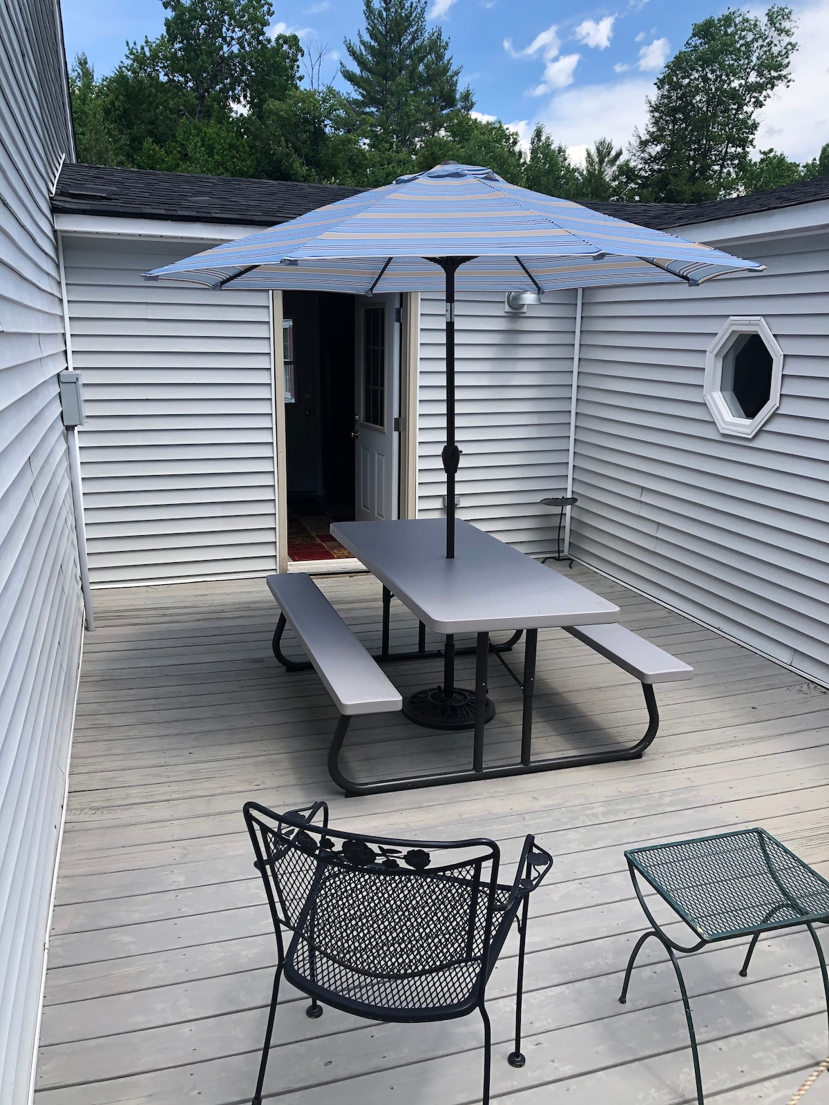 An outdoor space features a picnic table with an umbrella, surrounded by a deck. Two additional metal chairs are visible, providing seating options. The entrance to the home is accessible through a door that leads to the interior. The area is surrounded by trees.