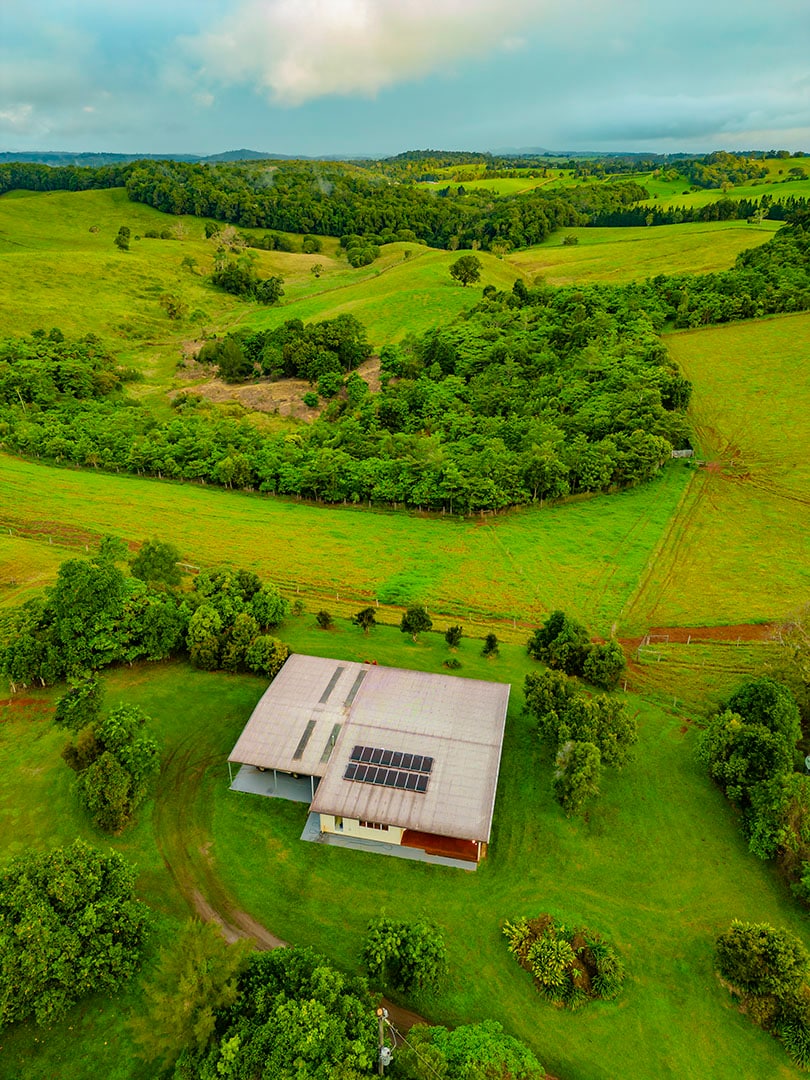 An aerial view captures the spacious home set amidst rolling green hills and lush forests. The building features a solar panel roof, and grassy fields extend in all directions, indicating a tranquil rural setting. The surrounding landscape showcases various shades of green.