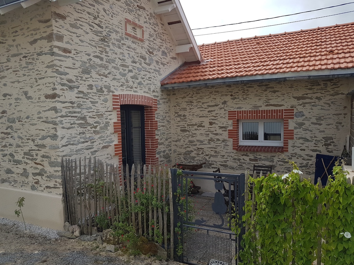The exterior of the house features natural stone walls complemented by a red-tiled roof. A fenced yard is visible, with a small garden area and a seating arrangement on a paved surface. Windows are adorned with decorative brick accents.