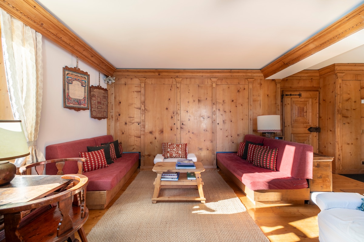 A spacious living area features warm wooden paneling and comfortable seating on red upholstery. A coffee table sits center, surrounded by books and decorative cushions. Natural light flows through large windows, offering a serene atmosphere for relaxation.