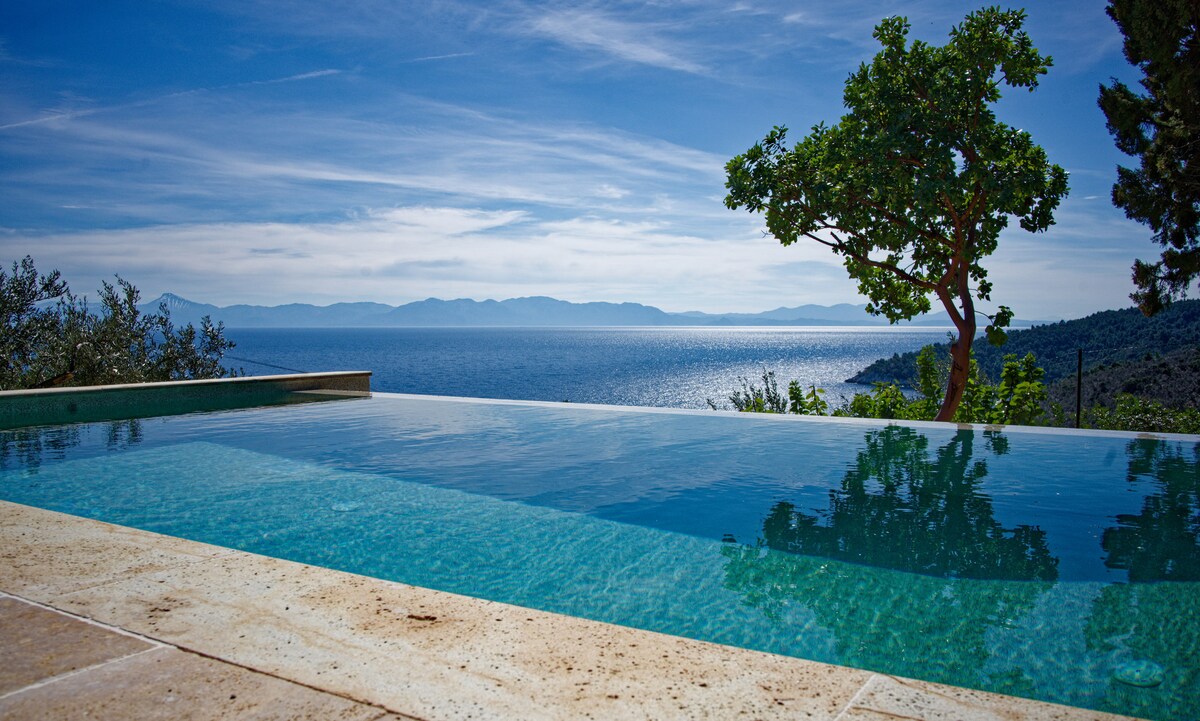 An infinity pool with clear water reflects the expansive sea views and the distant mountains. Olive and almond trees frame the pool area, enhancing the serene atmosphere. The sky above features soft, wispy clouds under natural light.