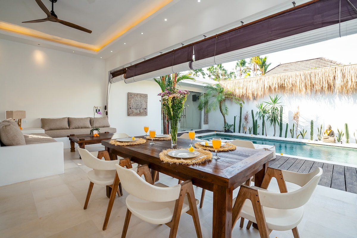 An open-air living area features a spacious wooden dining table set for breakfast, complemented by comfortable chairs. Soft furnishings in neutral tones are arranged on a cozy sofa in the corner, with tropical plants surrounding the private pool visible through large glass doors.