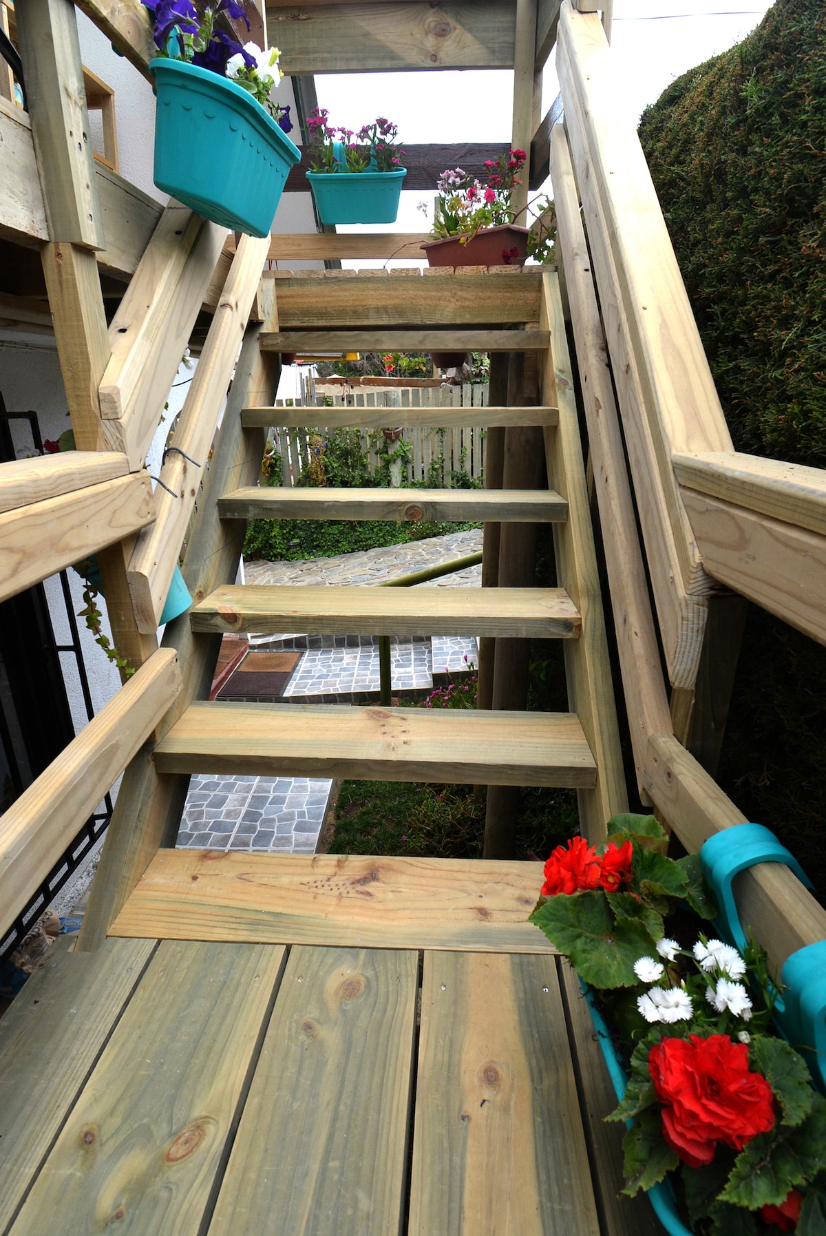Stairs made of wooden planks lead upward, framed by flower pots filled with vibrant blooms. A pathway with cobblestones is visible at the top, surrounded by greenery, creating a welcoming entrance to the private space.