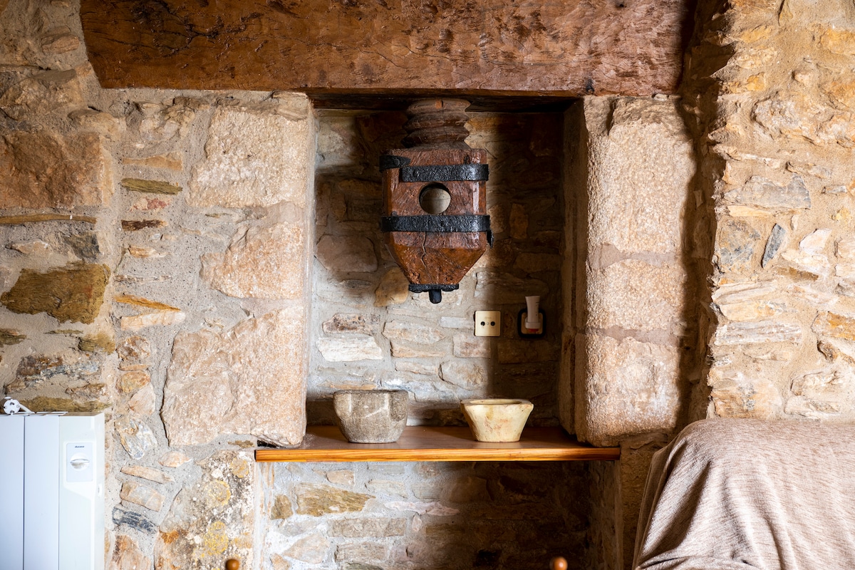 A traditional stone wall features a prominent wooden beam overhead. Below, a shelf displays two rustic bowls accompanied by an antique press, highlighting authentic Catalan design elements. The varied textures of the stones and wood contribute to the historical charm of the interior.