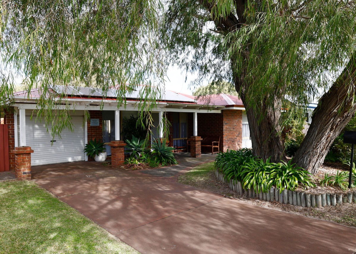 The exterior of the holiday home is framed by a large Peppermint tree, providing shade to the entrance. A paved driveway leads to the front door, flanked by landscaped gardens and low brick walls. A covered porch area includes a small chair for relaxation.