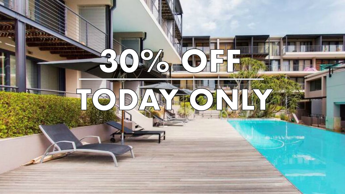 An outdoor area is showcased, featuring a wooden deck lined with lounge chairs. A shimmering pool is visible adjacent to the deck, with modern architecture in the background. Green foliage provides a touch of nature, complementing the well-maintained surroundings.