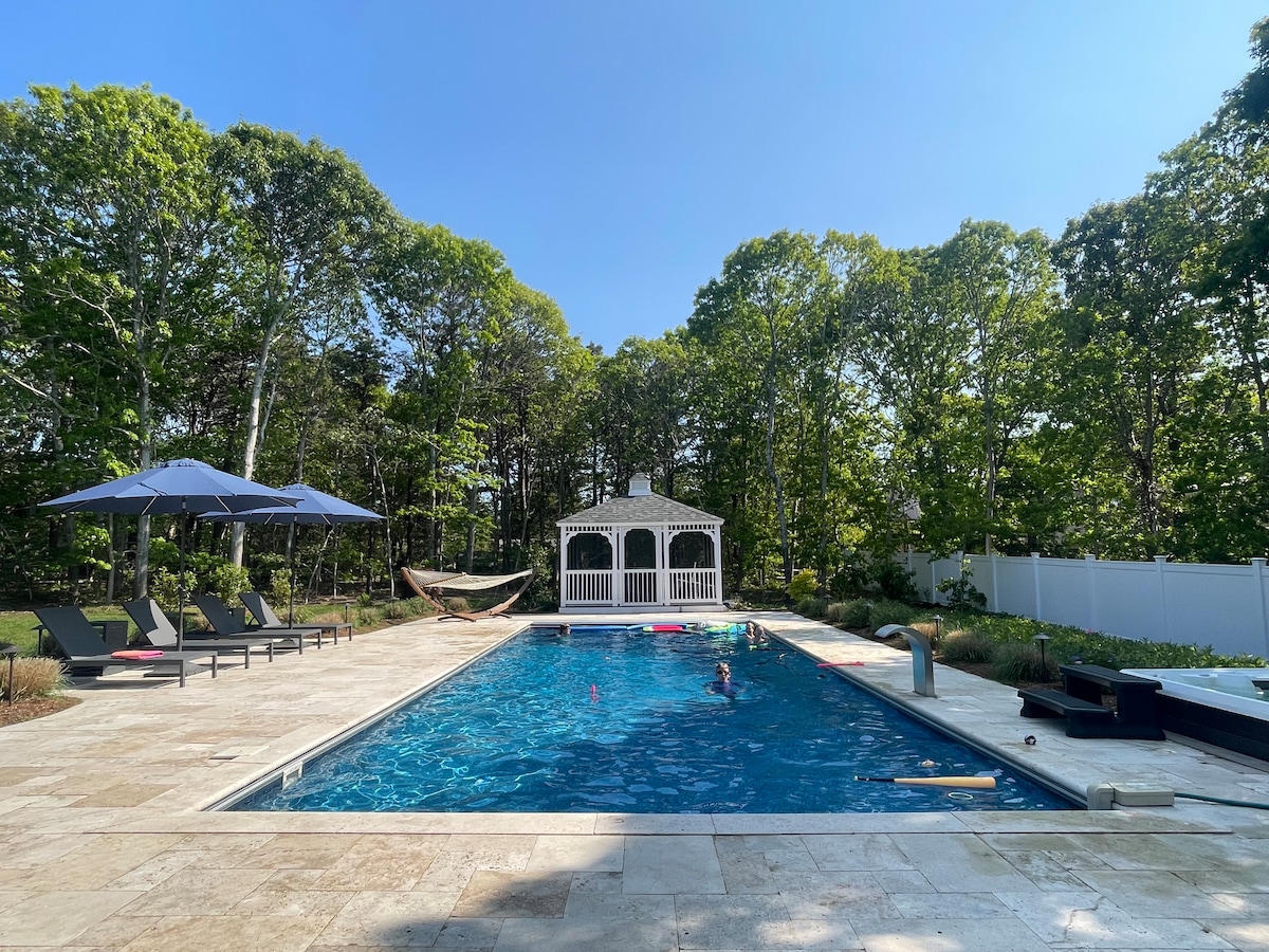 An expansive in-ground heated saltwater pool is surrounded by a spacious patio. A gazebo is situated nearby, framed by lush trees. Lounge chairs with umbrellas provide shaded seating, while a hammock offers a relaxing spot. The pool reflects the clear blue sky, creating a serene outdoor setting.