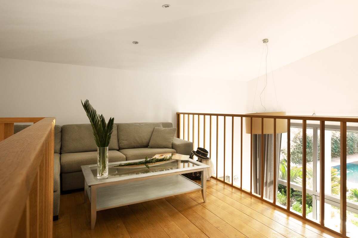 A bright loft area features a spacious light grey sofa paired with a sleek coffee table. Wooden flooring adds warmth, while natural light floods the space from nearby windows, providing a view of the outdoor pool area. Minimalist decor enhances the serene atmosphere.