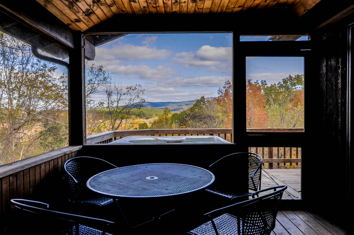 A covered balcony offers a view of the rolling hills and colorful foliage of the surrounding landscape. A black metal table and four matching chairs are positioned centrally, with a hot tub visible through the large glass windows, showcasing the serene outdoor setting.