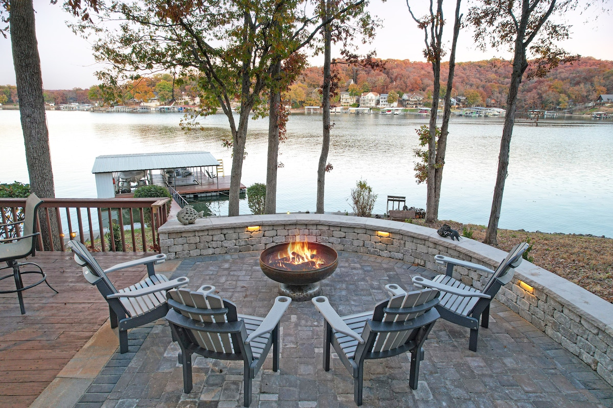 A stone patio is framed by natural landscaping, featuring a fire pit at its center surrounded by six outdoor chairs. The tranquil lake reflects trees with autumn foliage, providing a serene backdrop for gatherings.
