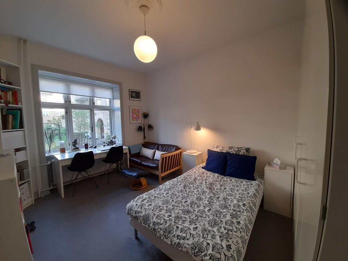A cozy bedroom features a spacious bed with a patterned duvet and two blue pillows. A seating area includes a dark sofa, a round side table, and two chairs, positioned by a large window that allows natural light to filter in.