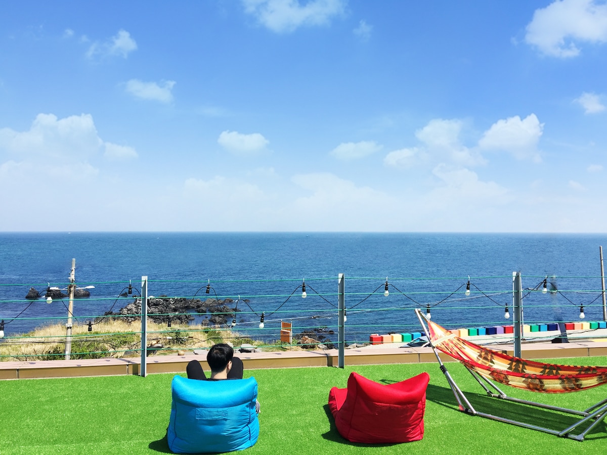 Brightly colored bean bags are positioned on a green lawn, facing a panoramic view of the ocean. A hammock is nearby, and string lights are visible, creating a relaxed atmosphere. The sky is clear blue with a few clouds, enhancing the scenic coastal setting.