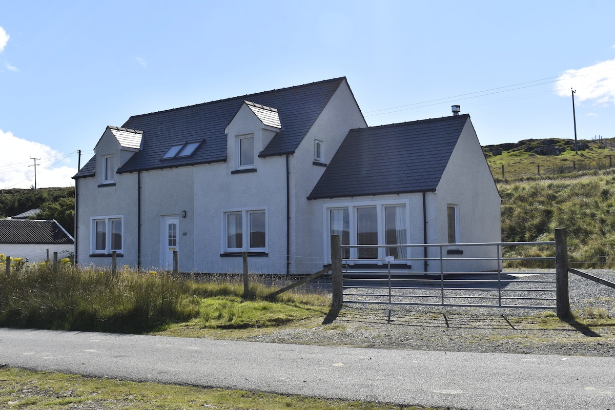 A modern cottage is shown from the exterior, featuring a light-colored facade with multiple windows. The structure sits amidst grassy terrain, with a gravel driveway leading to a gate. Countryside hills rise in the background under a clear blue sky.