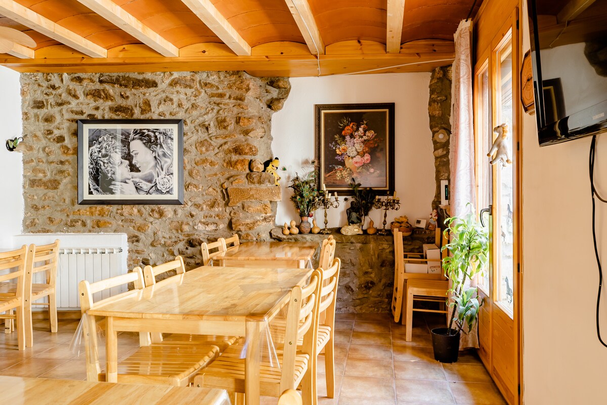 The dining area showcases light wooden tables accompanied by wooden chairs, arranged to create a spacious setting. A stone wall adds natural texture, while a floral artwork and framed photograph enhance the decor. Large windows allow natural light to fill the room.