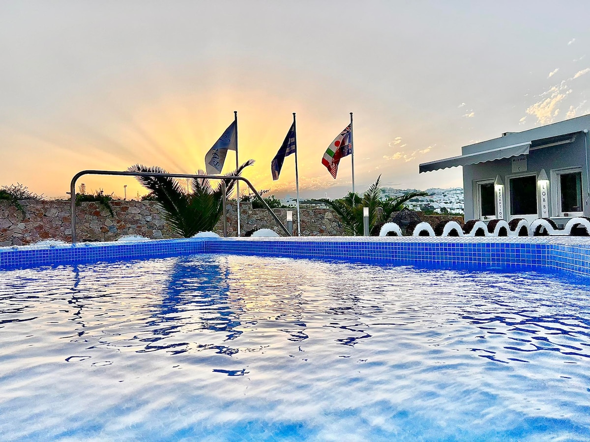 The image displays a tranquil swimming pool with a clear blue water surface reflecting the soft evening sky. Multiple flags are seen in the background, with a gentle sunset illuminating the scene. Lush plants are positioned near the pool's edge, enhancing the peaceful environment.