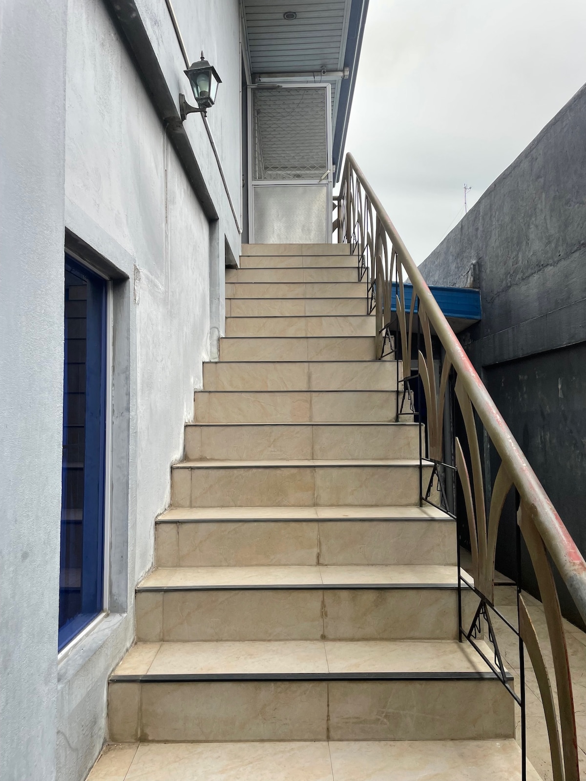 A set of outdoor stairs leads to the upper level, featuring a wrought iron railing and tiled steps. The walls are painted light gray, and a blue door is visible at the top. A light fixture hangs near the top of the stairs, providing illumination.