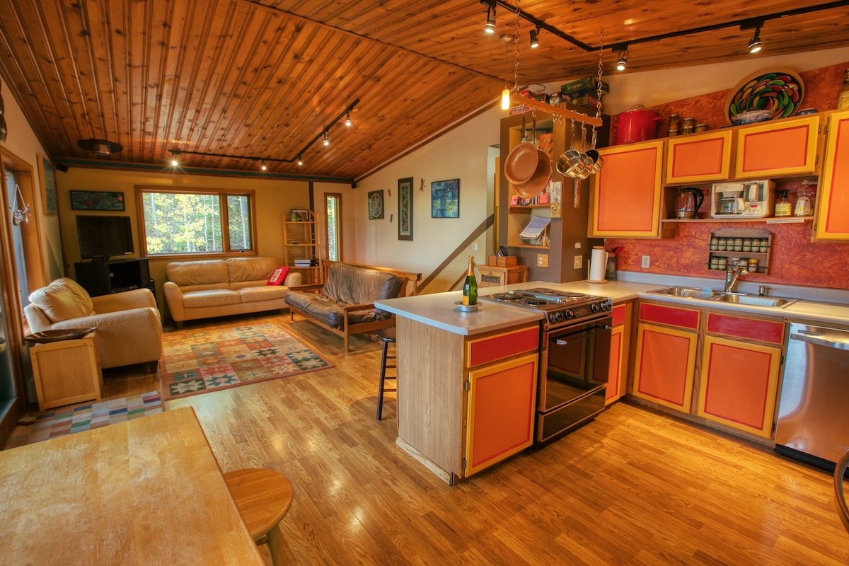 The kitchen area features wooden cabinetry with orange accents and open shelving. Stainless steel appliances, including a gas stove and refrigerator, are arranged for convenience. A spacious dining table and comfortable seating options are visible in the open layout, complemented by natural light from multiple windows.