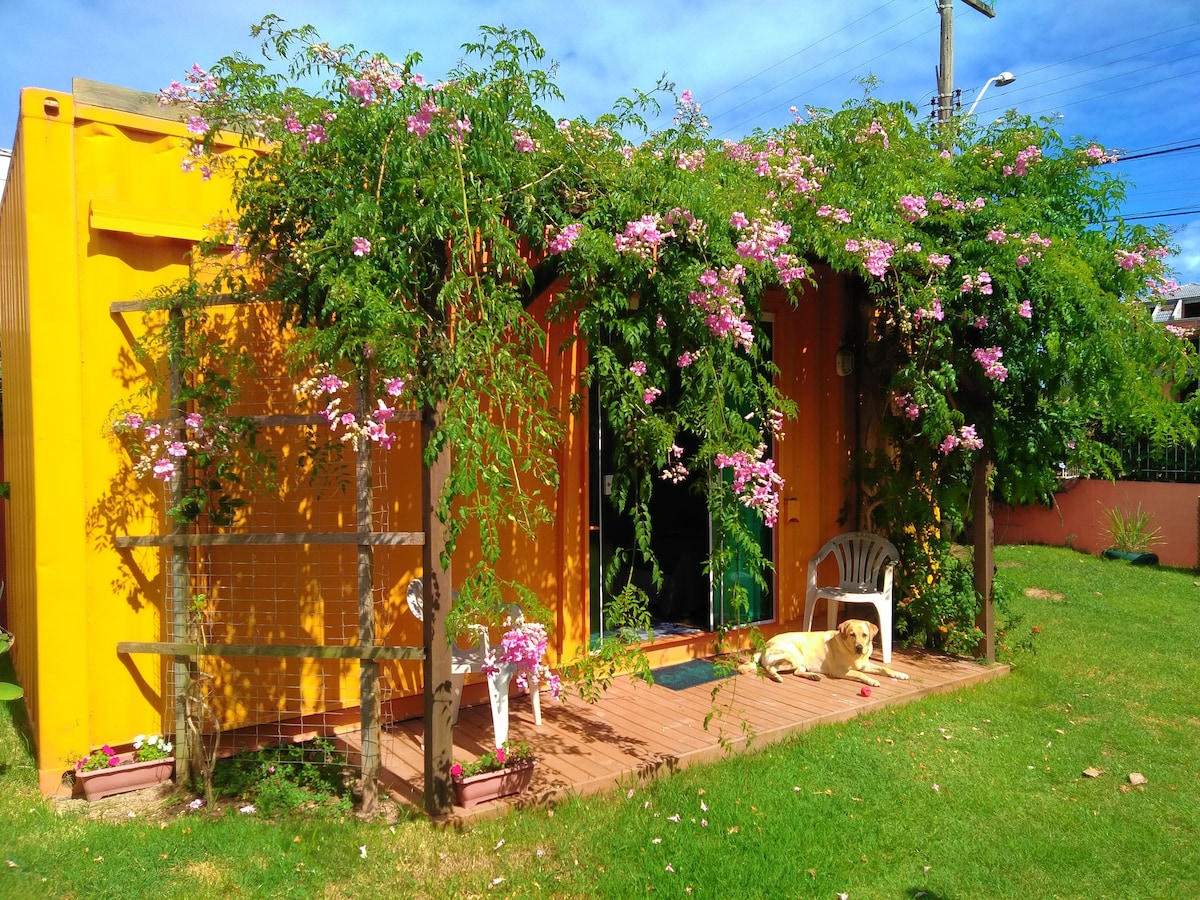 The exterior of the bright yellow 'Suíte Container' is adorned with lush flowering vines, creating a vibrant entrance. A cozy seating area features a white chair on a wooden deck, with a friendly dog resting nearby. The surrounding grassy space adds to the inviting atmosphere.